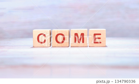 Four wooden blocks each display a letter together they spell COME in vibrant red against a pale, painted backdrop, conveying an invitation 134790336