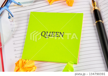 A bright green sticky note displays the word Queen on top of a planner. A pen rests beside it, and glasses and colorful paper origami add charm to the scene 134790391