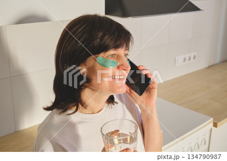 Mature Woman applying eye patches while talking on smartphone and drinking water in kitchen Mature Woman applying eye patches while talking on smartphone and drinking water in kitchen 134790987