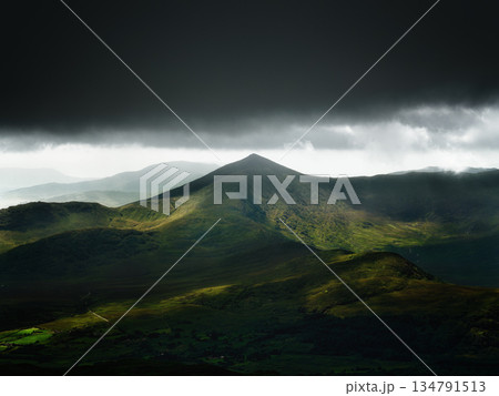 A stunning view of rugged mountain peaks in Ireland. Dark storm clouds loom above, while patches of sunlight illuminate the green valleys. A stunning view of rugged mountain peaks in Ireland. Dark storm clouds loom above, while patches of sunlight illuminate the green valleys. 134791513