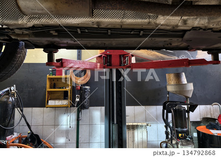 A car lift is positioned under a vehicle in an auto shop. Tools and equipment are visible in the area, indicating ongoing repairs and maintenance work A car lift is positioned under a vehicle in an auto shop. Tools and equipment are visible in the area, indicating ongoing repairs and maintenance work 134792868