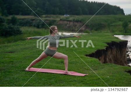 Caucasian woman doing yoga on the river bank. Warrior pose. Caucasian woman doing yoga on the river bank. Warrior pose. 134792987