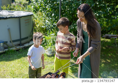 Caucasian woman grilling sausages on a barbecue with her two sons. Caucasian woman grilling sausages on a barbecue with her two sons. 134793147