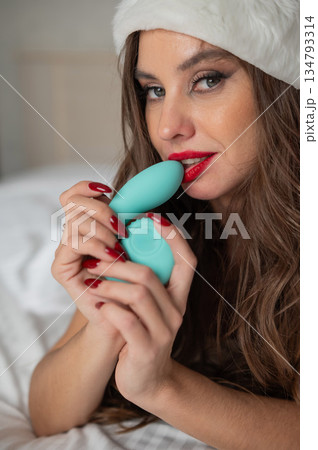 Caucasian woman in Santa Claus costume lying on bed holding curved sex toy.  134793314