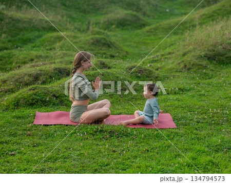 Caucasian woman doing yoga with her little son outdoors. Caucasian woman doing yoga with her little son outdoors. 134794573