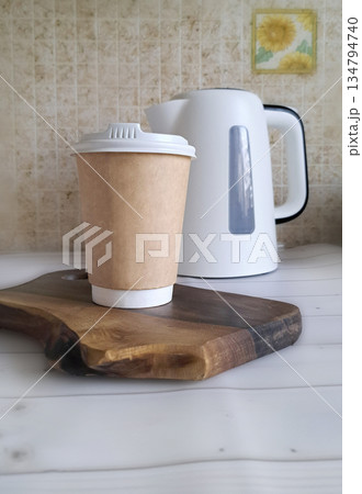 A paper coffee cup on a wooden board near an electric kettle 134794740