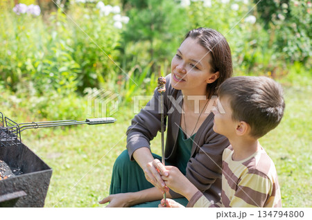 Caucasian woman grilling sausages on a barbecue with her son.  134794800