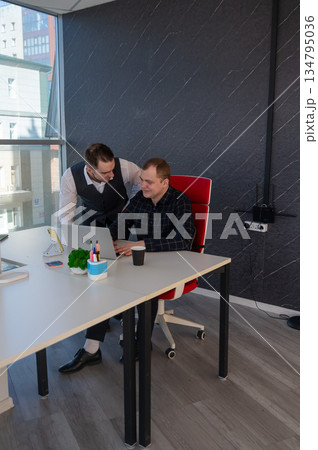 Caucasian man standing at colleague's desk discussing work. Caucasian man standing at colleague's desk discussing work. 134795036