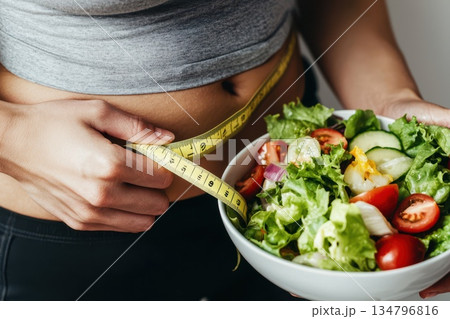 Person Measuring Waistline with Tape While Holding Bowl of Fresh Vegetables 134796816