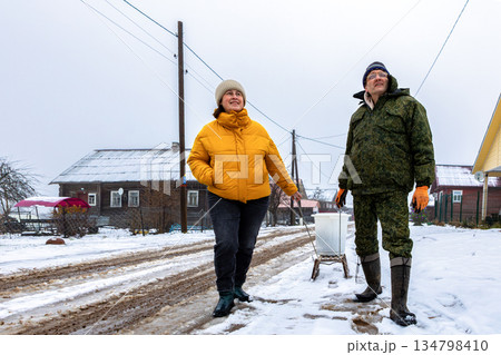 Trash hauling on sled in rural area in winter, mature couple works as team delivering garbage to large garbage dumpster. Trash hauling on sled in rural area in winter, mature couple works as team delivering garbage to large garbage dumpster. 134798410