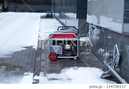 Portable red gasoline generator providing backup power to a modern building on a snowy winter day. 134798537