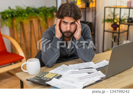 Indian man holds head over unpaid bills on table with calculator and laptop worried about money debt Indian man holds head over unpaid bills on table with calculator and laptop worried about money debt 134799242