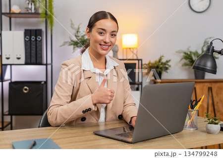 Young happy businesswoman shows thumbs up to camera confirms task complete on laptop smiles with joy 134799248