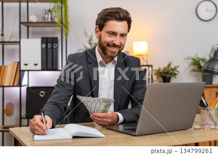 Middle-aged businessman counts cash dollars records budget on laptop writes notes in notebook plans 134799261