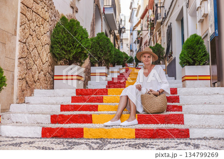 Young woman in a white dress sitting on the colourful Spanish Steps in the old town of Calpe, Spain, a well-known landmark painted in red and yellow  134799269