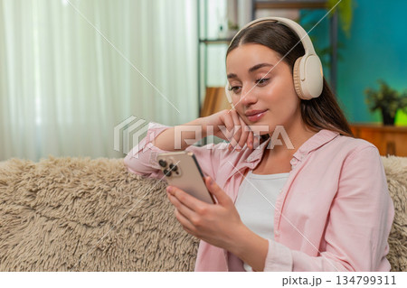Young woman listening to podcast on sofa wearing headphones nodding while writing insightful notes 134799311