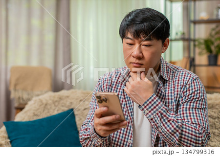 Asian man reading important message on smartphone, pausing scroll, tense thoughtful reaction on sofa Asian man reading important message on smartphone, pausing scroll, tense thoughtful reaction on sofa 134799316
