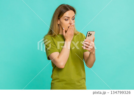 Adult woman scared looking at smartphone screen covering mouth reacting to shocking video bad news 134799326
