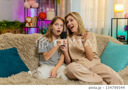 Child girl kid with mother singing karaoke with microphone together at home on living room cozy sofa 134799344