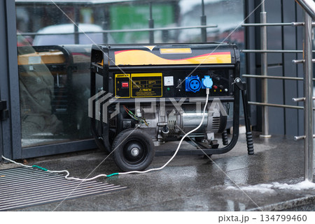 Portable black gasoline generator with connected power cables operating outside a store on a wet winter day 134799460