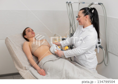 Caucasian woman receives abdomen vacuum massage from cosmetologist using LPG device in beauty clinic 134799504