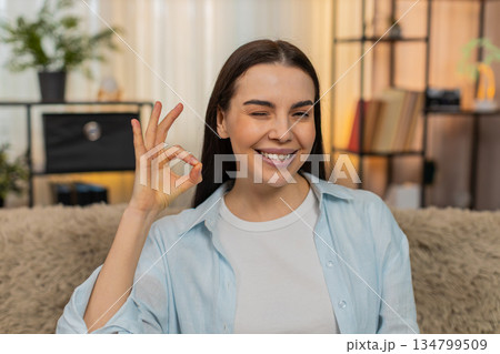 Young woman sitting on sofa happily smiling confidently showing OK hand sign gesture positive mood 134799509