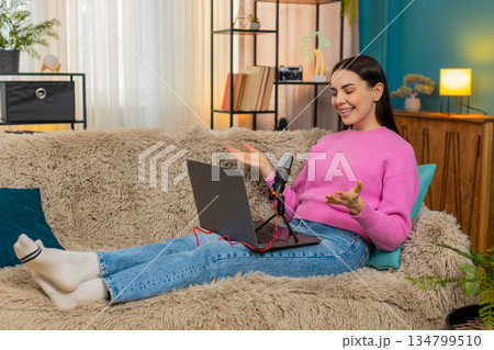 Young woman sitting on sofa recording podcast speaking seriously into microphone with focused look Young woman sitting on sofa recording podcast speaking seriously into microphone with focused look 134799510