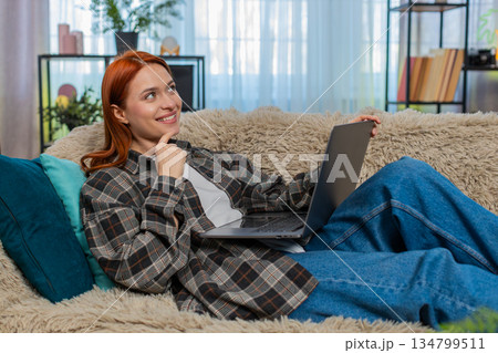 Woman sitting on sofa shopping online with laptop choosing dress touching chin in thoughtful pose Woman sitting on sofa shopping online with laptop choosing dress touching chin in thoughtful pose 134799511