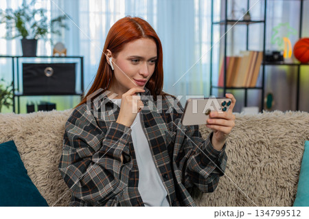 Woman sitting on sofa wearing headphones watching video on smartphone enjoying social media content Woman sitting on sofa wearing headphones watching video on smartphone enjoying social media content 134799512