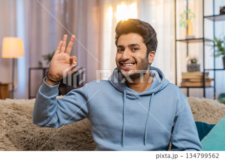 Happy Indian man showing OK gesture and smiling confidently while sitting on sofa at home room 134799562