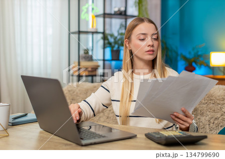 Caucasian woman managing tax declaration at home on sofa table, holding documents, calculator laptop Caucasian woman managing tax declaration at home on sofa table, holding documents, calculator laptop 134799690