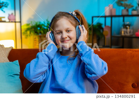 Happy child girl in wireless headphones relaxing sitting on sofa at home listening favorite music 134800291
