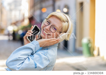 Carefree young woman in glasses listening music via wireless headphones while walking on city street Carefree young woman in glasses listening music via wireless headphones while walking on city street 134800304