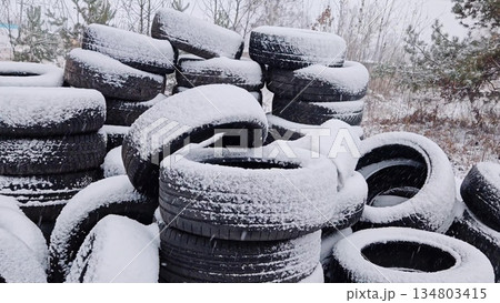 Stacking used car tires covered with fresh snow outdoors. Piles of old automobile tires accumulating snow in winter storage yard during snowfall 134803415
