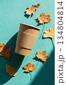 Eco coffee cup and autumn leaf against a teal background. Autumn paper coffee cup and fall leaves on vibrant teal background. 134804814