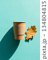 Eco coffee cup and autumn leaf against a teal background. Kraft coffee cup and dry autumn oak leaf flat lay on turquoise background. 134804815
