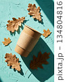 Eco coffee cup and autumn leaf against a teal background. Disposable coffee cup and autumn dry leaves on bright textured teal background. 134804816