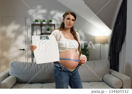 Pregnant woman holding scale and measuring her belly with tape measure indoors with soft lighting Pregnant woman holding scale and measuring her belly with tape measure indoors with soft lighting 134805532