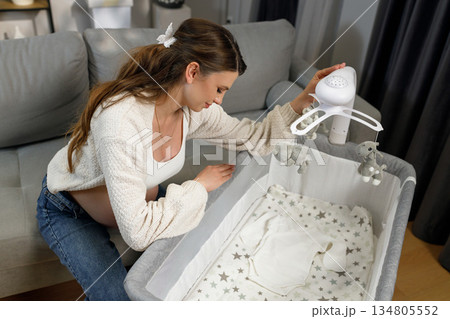 Expecting mother prepares nursery by attaching mobile to baby crib, anticipating newborn arrival with gentle care and excitement Expecting mother prepares nursery by attaching mobile to baby crib, anticipating newborn arrival with gentle care and excitement 134805552