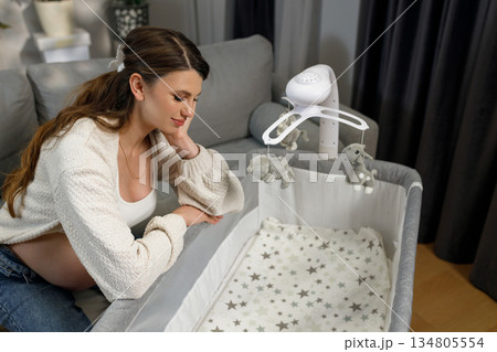 Expecting mother sits on sofa, looking thoughtfully at baby crib with mobile, anticipating motherhood and nursery setup Expecting mother sits on sofa, looking thoughtfully at baby crib with mobile, anticipating motherhood and nursery setup 134805554