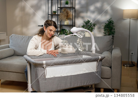 Young mother gazes lovingly at a crib with a mobile, creating a serene nursery atmosphere in a modern living room Young mother gazes lovingly at a crib with a mobile, creating a serene nursery atmosphere in a modern living room 134805558