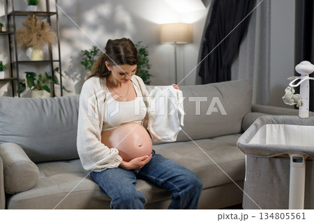 Expecting mother sits on sofa holding tiny white baby onesie, preparing for newborn arrival with crib nearby Expecting mother sits on sofa holding tiny white baby onesie, preparing for newborn arrival with crib nearby 134805561