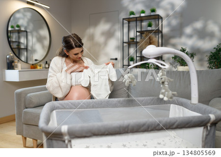 Expecting mother sits on sofa holding tiny white onesie near crib, preparing nursery with anticipation and joy 134805569
