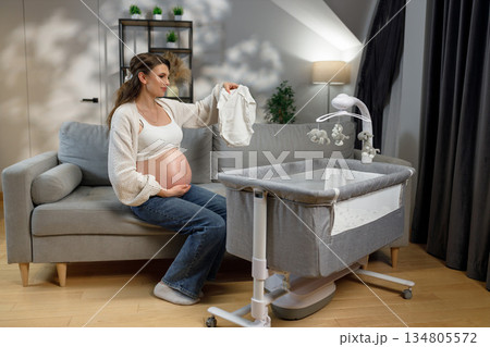 Expecting mother sits on sofa holding baby garment near bedside bassinet, preparing nursery room for newborn arrival Expecting mother sits on sofa holding baby garment near bedside bassinet, preparing nursery room for newborn arrival 134805572