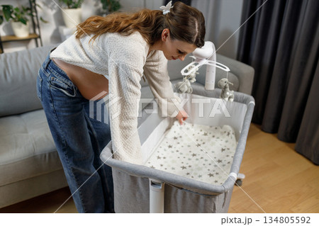 Pregnant woman arranging bedding in portable baby crib, preparing nursery for newborn arrival Pregnant woman arranging bedding in portable baby crib, preparing nursery for newborn arrival 134805592