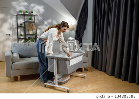 Pregnant woman arranging bedding in a bedside bassinet next to sofa in living room with plants and curtains Pregnant woman arranging bedding in a bedside bassinet next to sofa in living room with plants and curtains 134805595