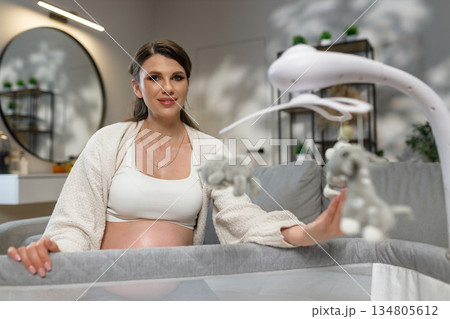 Pregnant woman sitting near crib with baby mobile, anticipating motherhood and nursery preparation 134805612