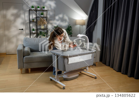 Woman leans over bedside bassinet, adjusting hanging toys on mobile, preparing nursery for infant Woman leans over bedside bassinet, adjusting hanging toys on mobile, preparing nursery for infant 134805622