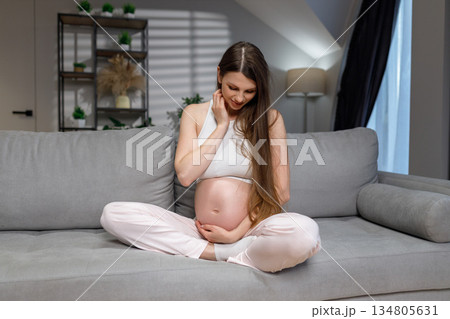 Young expectant mother sits cross-legged on couch, cradling her baby bump with gentle smile, indoors Young expectant mother sits cross-legged on couch, cradling her baby bump with gentle smile, indoors 134805631