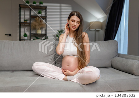 Young expectant mother sits cross-legged on a comfortable couch, cradling her belly with a gentle smile Young expectant mother sits cross-legged on a comfortable couch, cradling her belly with a gentle smile 134805632